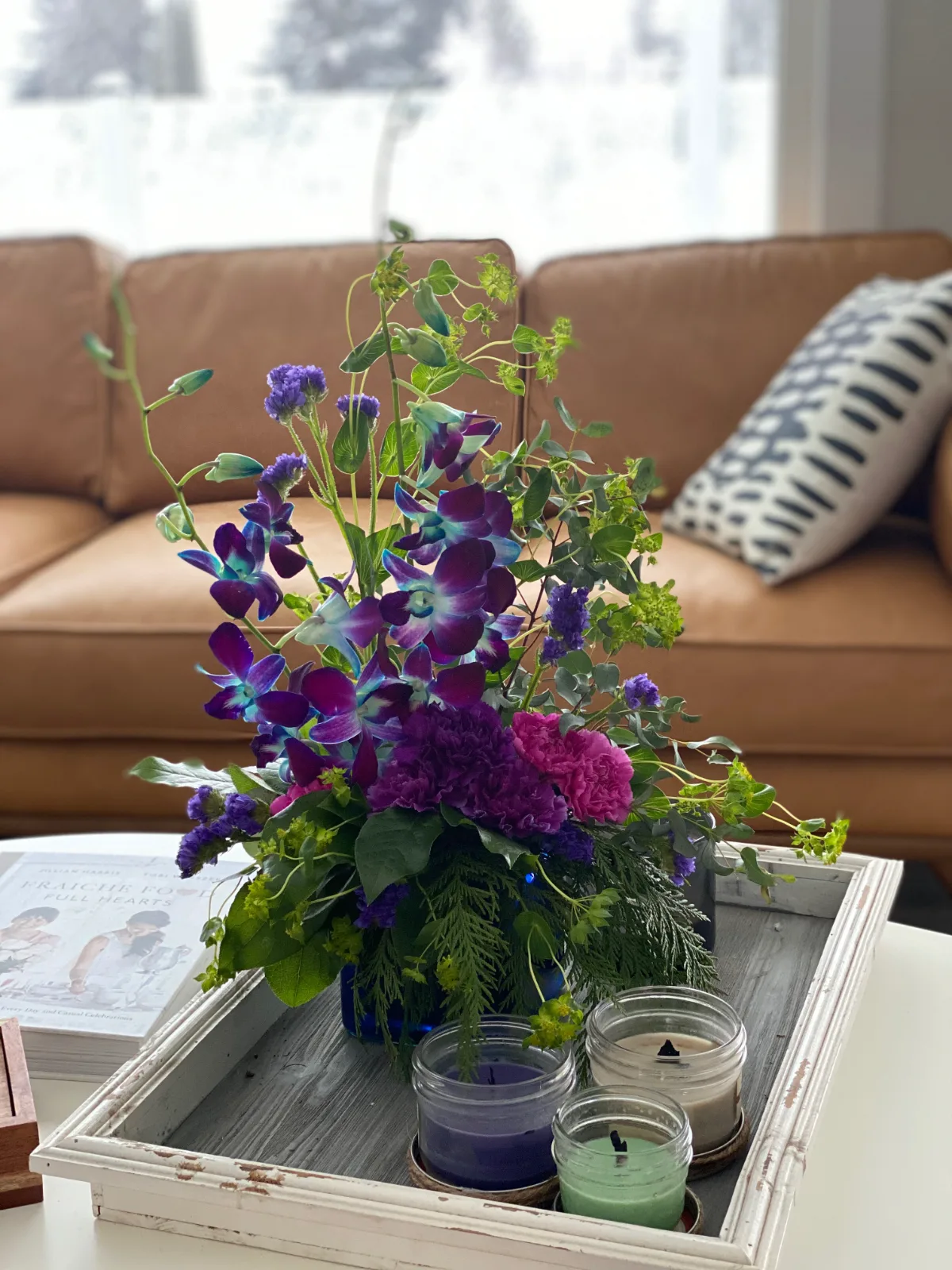Purple orchids and carnations with trailing greenery in a low vase on a wooden tray beside two candles, on a living room table.