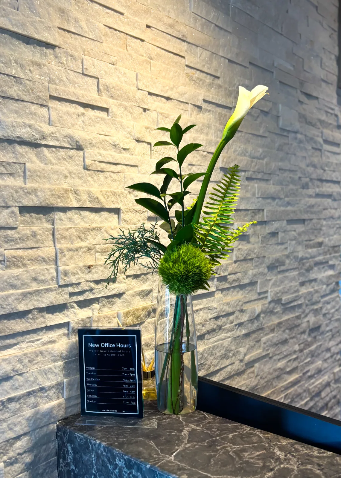 A white calla lily, a green chrysanthemum and fern in a slim glass vase on a stone ledge beside an office hours sign.