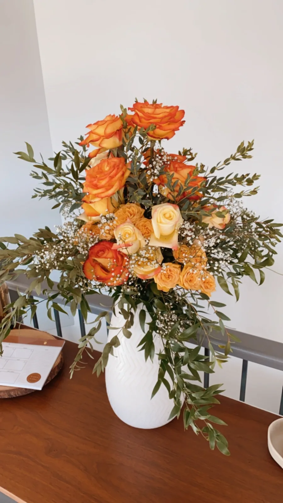 Orange roses and peach blooms with eucalyptus in a white vase on a wooden sideboard.