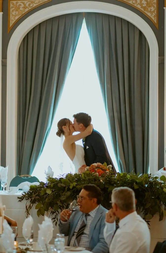 The couple kissing at the head table under a tall arched window, with a long garland of flowers along the table.