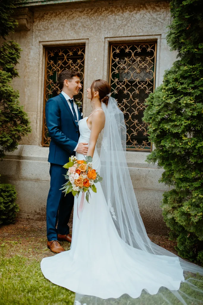 The bride and groom by an old stone wall with leaded windows, the bride holding a bouquet of orange and peach roses.