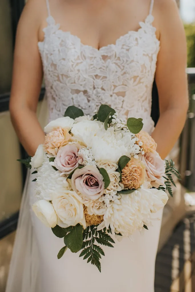 A bride in a lace gown holding a bouquet of white and blush roses, cream ranunculus and peach blooms with fern.