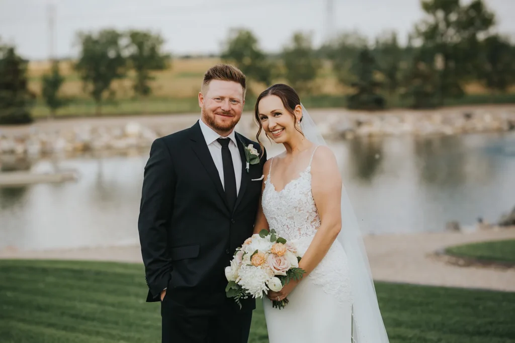 The bride and groom by a pond, the bride holding a bouquet of white and blush flowers.
