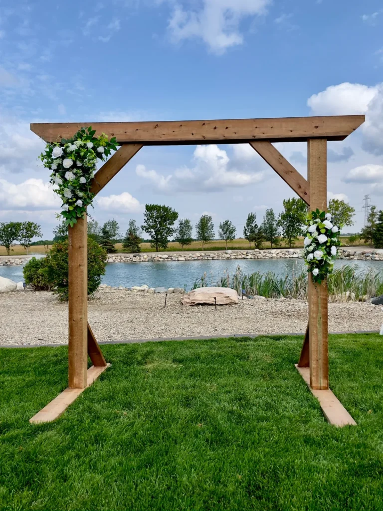 A wooden ceremony arch dressed with white flowers and greenery on a lawn by a pond.