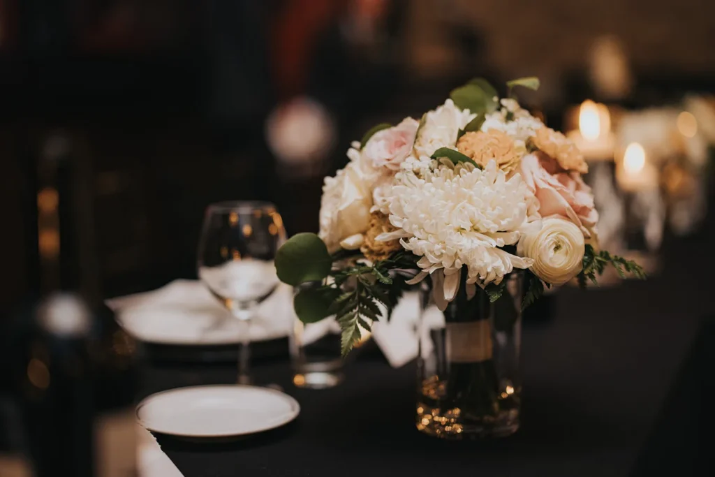 A low arrangement of white and blush roses and ranunculus on a dark table by candlelight.