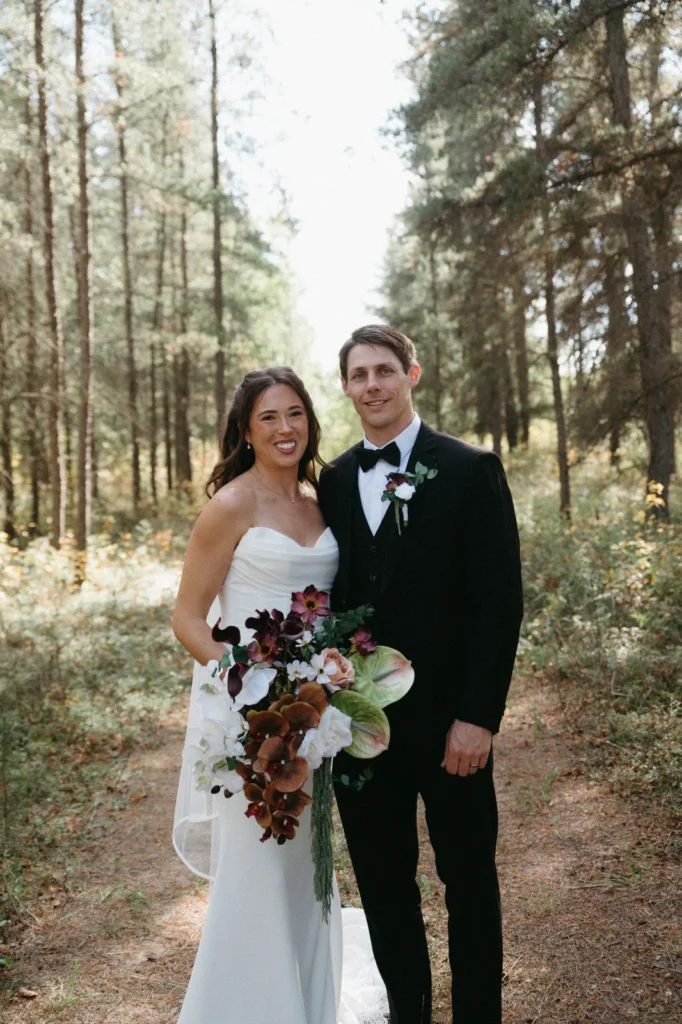 The bride and groom on a path through tall pines, the bride holding a bouquet of burgundy, white and rust flowers.