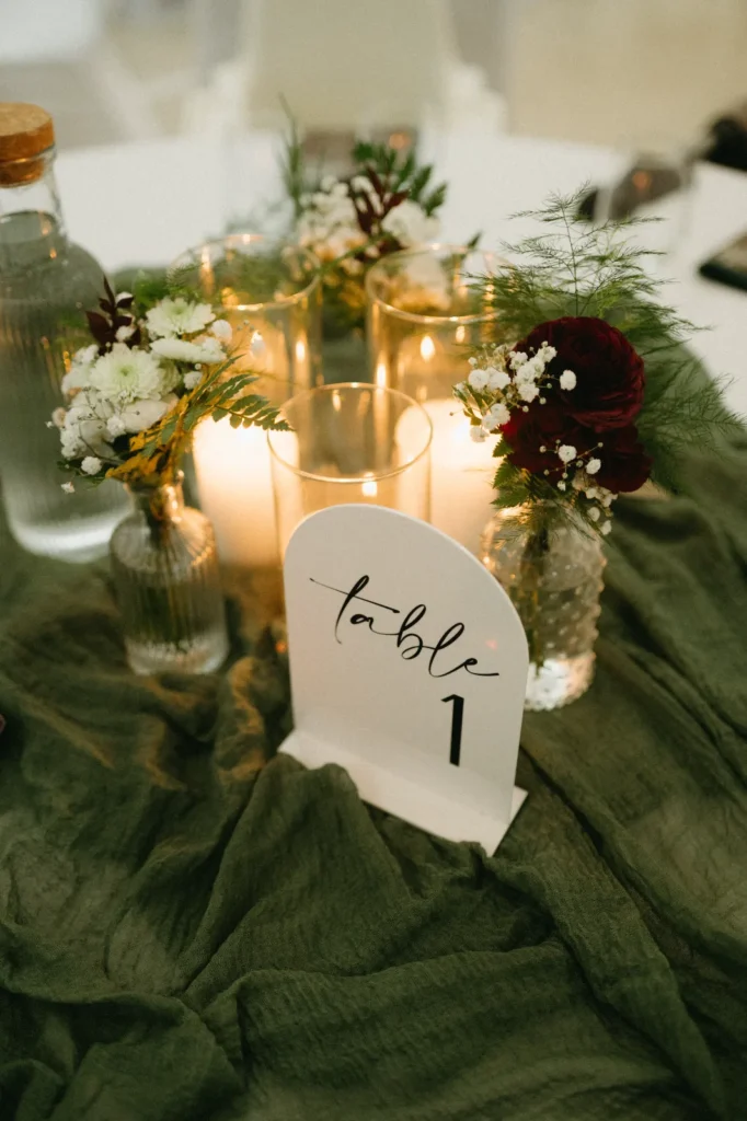 Small bottles of white and burgundy flowers with candles and a table number on a green cloth.