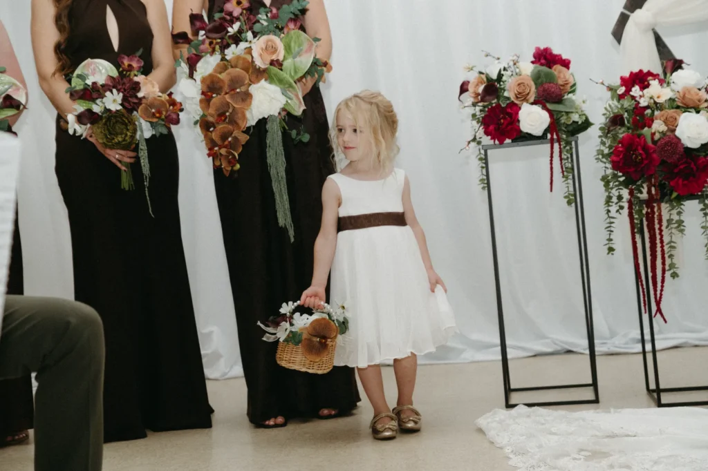 A flower girl with a basket beside the bridesmaids, with flowers on tall stands behind.
