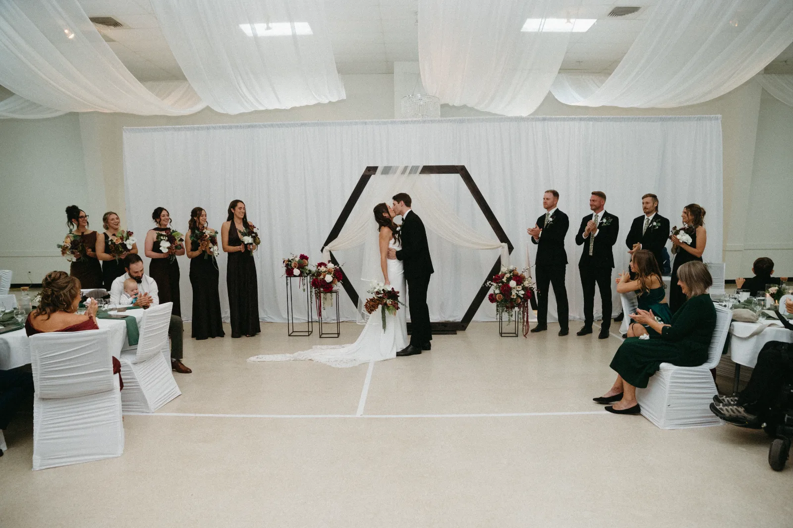 The ceremony in a draped hall, the couple under a hexagon arch with flowers on stands either side.