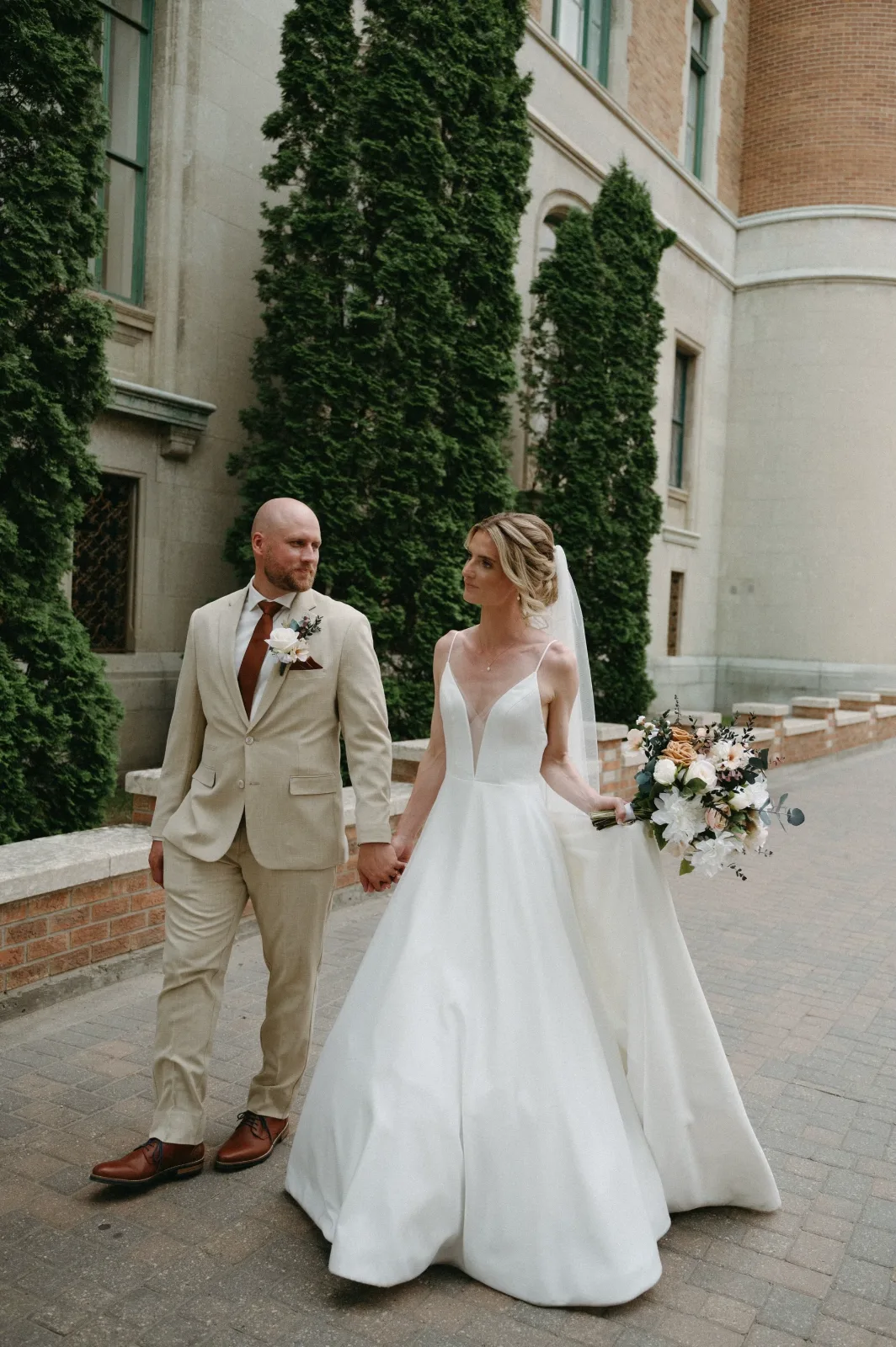 The bride and groom walking hand in hand between stone university buildings, the bride holding a peach and white bouquet.
