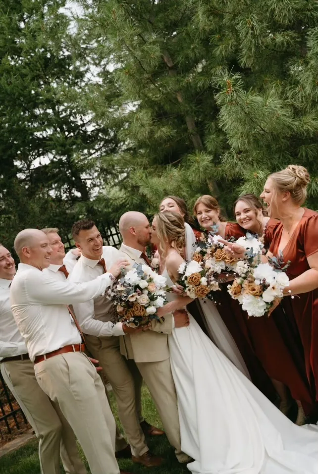 The wedding party laughing together under the trees, the bridesmaids holding bouquets of peach and cream flowers.