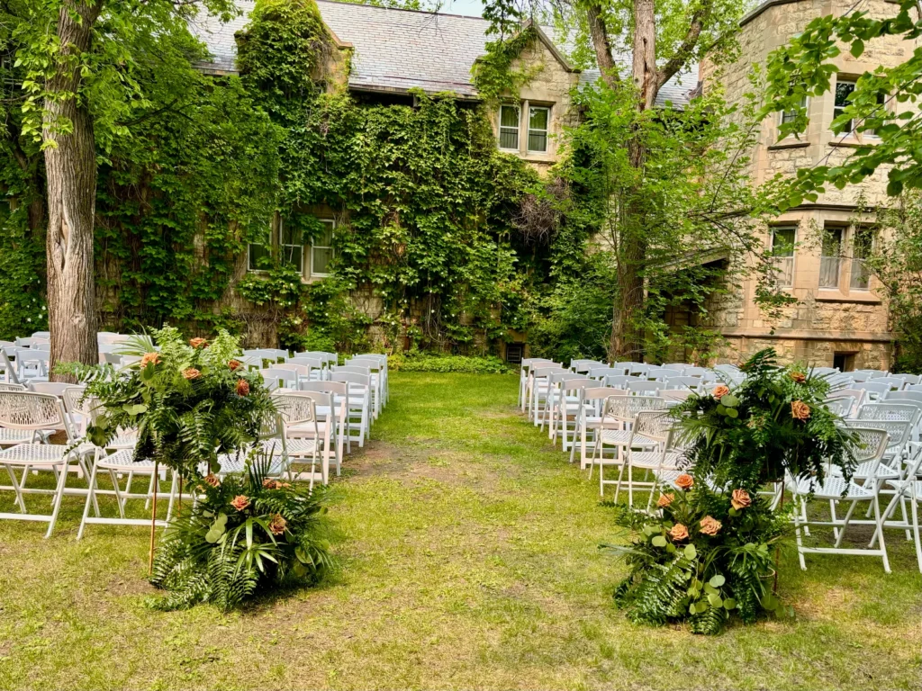White chairs in a green courtyard with an ivy-covered stone building behind, and flower arrangements on the ground at the end of each row.