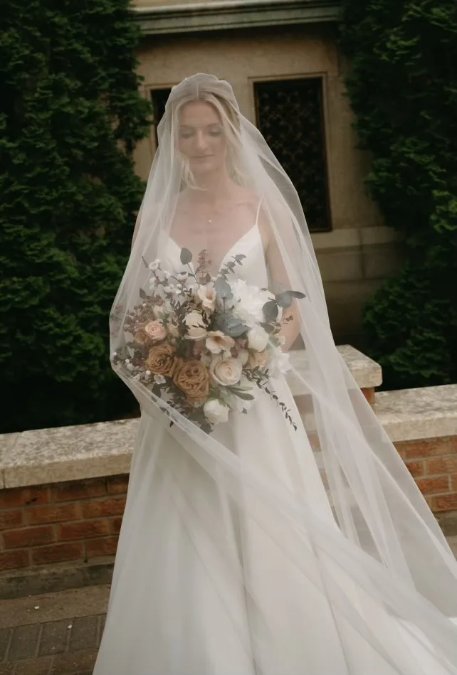 The bride in a long veil looking down at her bouquet of peach roses and cream flowers.
