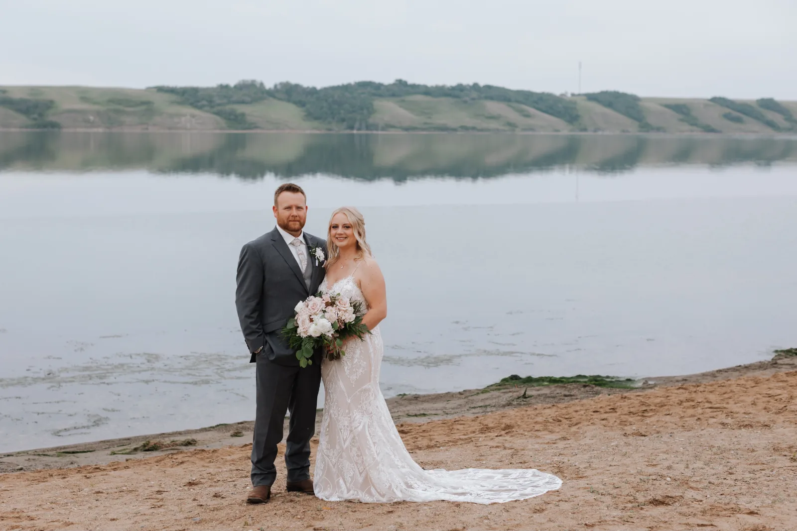 The bride and groom on a sandy beach with the lake and hills behind them, the bride holding a bouquet of blush and white flowers.