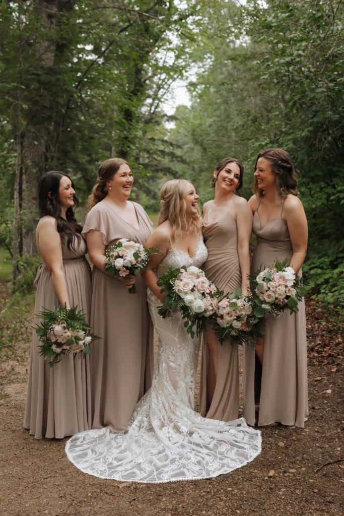 The bride laughing with four bridesmaids in taupe dresses under the trees, all holding bouquets of blush and white flowers.