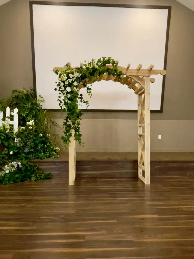 A wooden arch draped with greenery and small white flowers, with more greenery gathered at its foot, on a wooden floor.