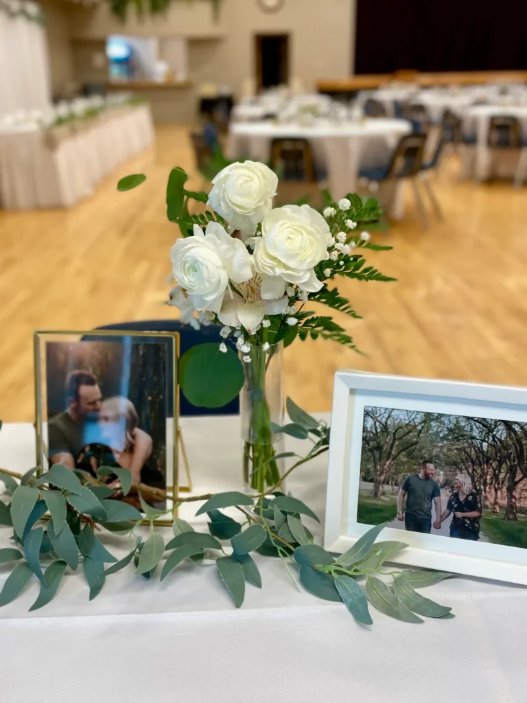 White roses and baby's breath in a small glass vase between framed photographs of the couple, with eucalyptus laid along the table.