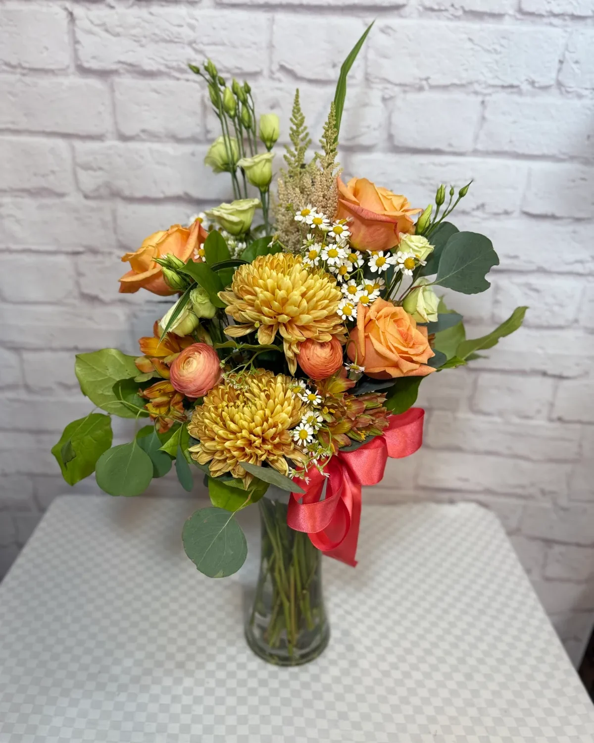 Orange roses, golden chrysanthemums and ranunculus with chamomile and eucalyptus in a glass vase tied with a coral ribbon.