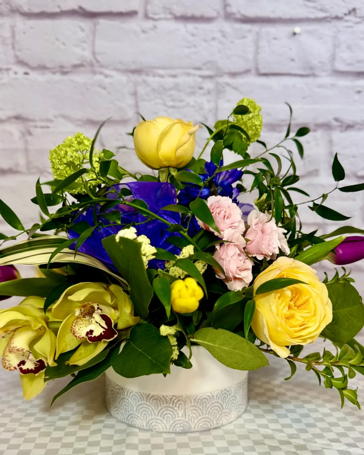 Yellow garden roses, green orchids, purple tulips and pink carnations in a low white container.