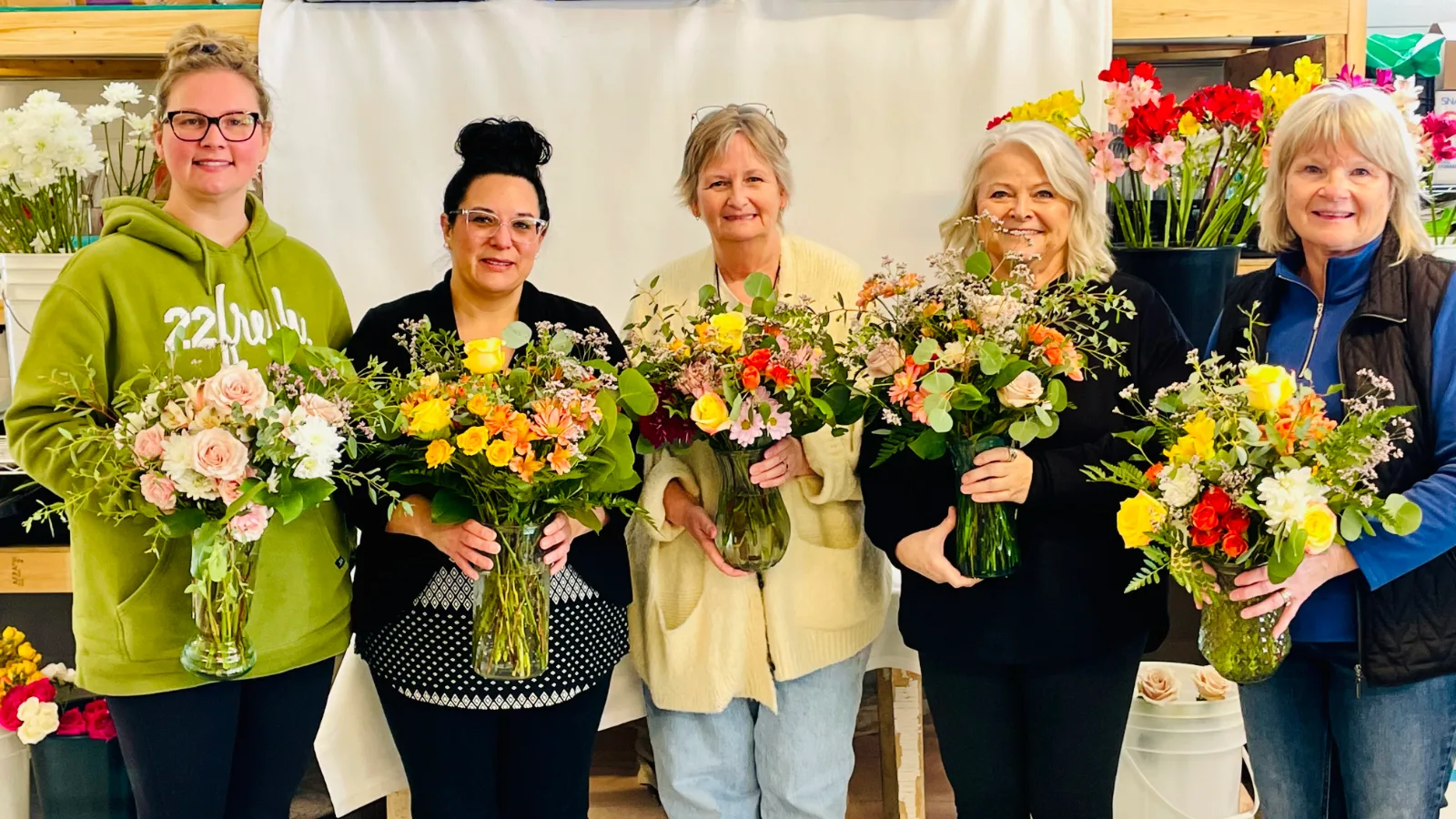 Five guests in Anne's studio, each holding the vase arrangement she made: pink roses, yellow and orange roses and mixed garden flowers, with buckets of stems on the shelves behind.