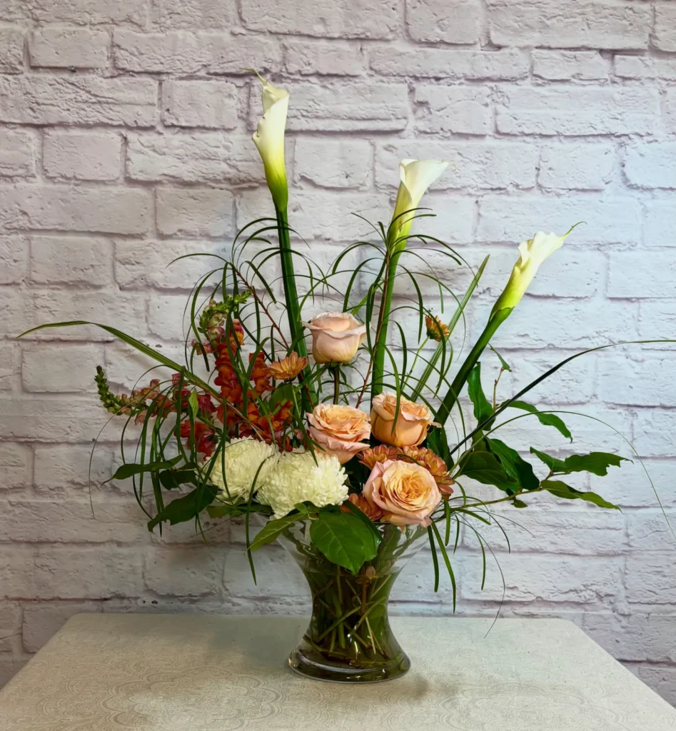 Three white calla lilies, peach roses, white chrysanthemum and orange snapdragons with grasses in a glass vase, against a white brick wall.