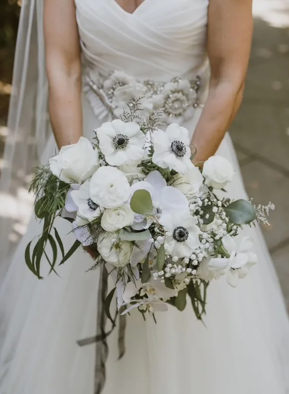 A bride in a white gown holding a bouquet of white anemones, ranunculus and baby's breath with eucalyptus and fern.