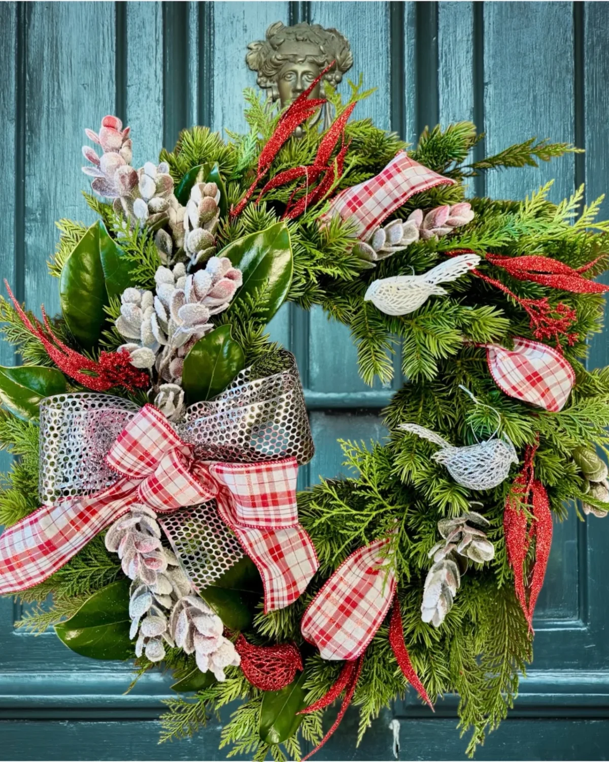 A Christmas wreath of fir and cedar with magnolia leaves, frosted cones, red glitter sprays, white and silver birds and a red and white plaid bow, on a teal front door.